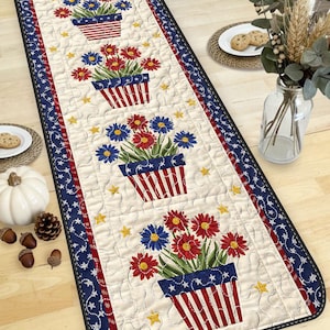 May include: Quilted table runner with a patriotic theme. Features red, white, and blue floral arrangements in striped pots, surrounded by stars. The runner has a cream background and a navy blue border.