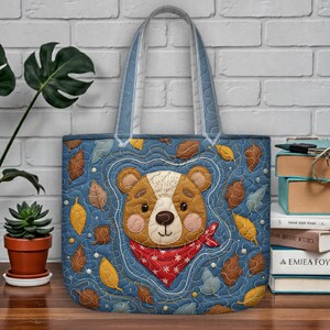Puede incluir: Un bolso tote acolchado con un diseño de cara de oso. El bolso presenta un fondo azul con hojas de otoño y una cara de oso con un pañuelo rojo. Las asas son de color azul claro.
