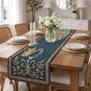 Op de afbeelding: Marineblauwe tafelloper met een gedetailleerd bloemen- en vogelontwerp. De loper heeft een gewatteerd patroon en een bruine rand. De tafel is gedekt met witte borden, kaarsen en een vaas met witte bloemen.