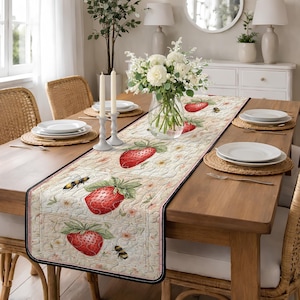 Puede incluir: Camino de mesa acolchado con diseño de fresas y abejas. El camino tiene un fondo crema con fresas rojas y hojas verdes. El borde es negro con un fino ribete rosa. Sobre una mesa de madera.