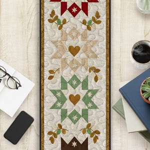 May include: A quilted table runner with a repeating star and heart pattern in red, tan, green, and brown. The runner has a gold border and is set against a light background. The design includes small star and leaf accents.