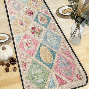 May include: A rectangular table runner with a quilted design featuring pastel-colored Easter egg shapes and floral patterns. The runner has a black border and is placed on a wooden table. The color palette includes pink, blue, yellow, and green.