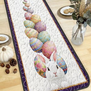 May include: Easter table runner with a white rabbit and colourful Easter eggs. The runner has a white quilted background, a purple border, and gold trim. The eggs are decorated with floral and geometric patterns.