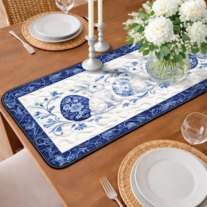 May include: A blue and white quilted table runner with a floral and bunny design. The runner features a dark blue border and a white background with blue floral and bunny motifs. The runner is on a wooden table, with plates, silverware, and a vase of white flowers.