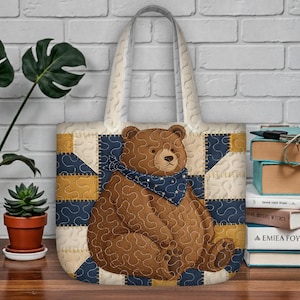 Op de afbeelding: Een gewatteerde draagtas met een bruine beer met een blauwe sjaal versierd met witte sterren. De tas heeft een patchwork ontwerp in crème, marineblauw en goud. De handvatten zijn crèmekleurig.