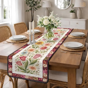 May include: A floral table runner with a white background and a colorful pattern of flowers, including red tulips, yellow sunflowers, and blue daisies. The runner has a dark border and is placed on a wooden dining table.