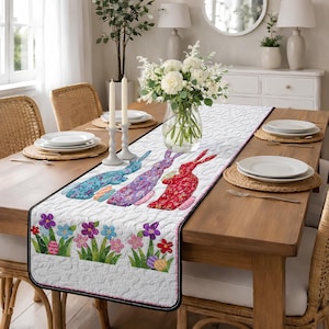 May include: A white Easter table runner with a black border, featuring three colorful bunny rabbits and floral and egg designs. The runner is placed on a wooden table set for a meal, with plates, candles, and a flower arrangement.