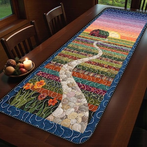 Könnte beinhalten: Gesteppter Tischläufer mit Landschaftsdesign. Der Läufer zeigt einen gewundenen Steinweg, der zu einem Sonnenuntergang führt, mit bunten Feldern und Blumen. Die Ränder sind dunkelblau mit weißen Nähten.