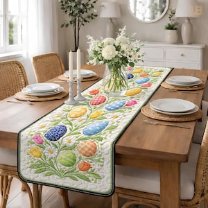 May include: A cream-colored Easter table runner with a floral and egg design. The runner features colorful eggs, tulips, and daisies. The edges are trimmed with a black border. The runner is placed on a wooden table with place settings and a vase of flowers.