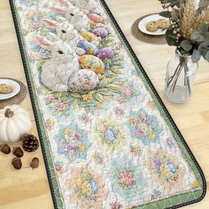 May include: Quilted Easter table runner featuring white bunnies, colorful Easter eggs, and floral patterns. The runner has a green border and a black stitched edge. The design is in pastel colors, creating a festive and spring-like atmosphere.