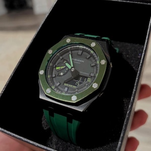 May include: A black and green G-Shock Casio watch in a black box. The watch has a green bezel and strap, with a dark grey face. The watch face displays the time and other functions. The watch is a stylish accessory.