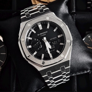 Puede incluir: Un reloj G-Shock Casio plateado con esfera negra y pantalla digital que muestra la hora. El reloj tiene una correa de acero inoxidable y una esfera redonda con detalles plateados. El reloj se muestra sobre una superficie negra.