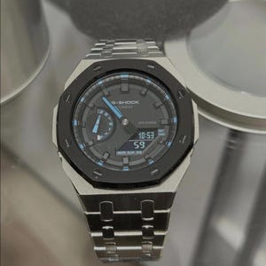 May include: A silver and black G-Shock Casio watch with a digital display showing the time. The watch has a black bezel and a silver strap. The watch face is dark grey with blue accents. The watch is displayed on a reflective surface.