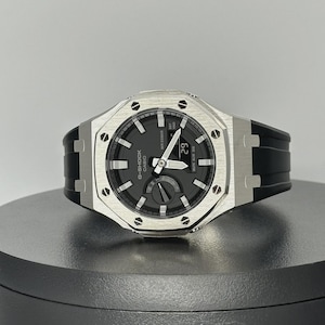 May include: A G-Shock Casio watch with a black face and a silver-tone bezel. The watch has a black rubber strap and displays the date as '29'. The watch is water resistant.