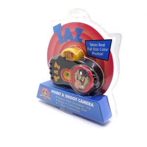 May include: A black and red point and shoot camera featuring Taz from Looney Tunes. The camera is packaged in a clear plastic clamshell with a red and yellow background. The packaging includes the text "Takes Real Full Size Color Photos!" and "Focus Free Outdoor 110, Drop-in Cartridge Film, 1 Year Guarantee".
