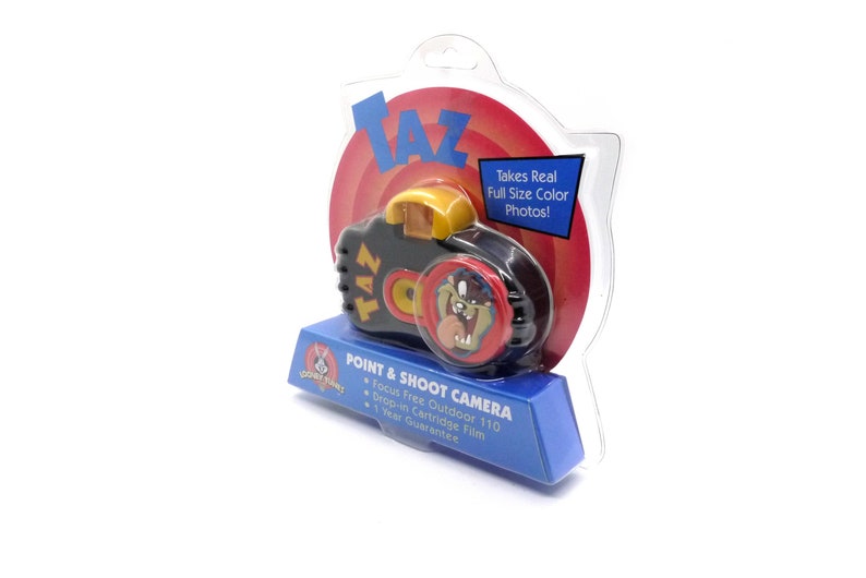 May include: A black and red point and shoot camera featuring Taz from Looney Tunes. The camera is packaged in a clear plastic clamshell with a red and yellow background. The packaging includes the text "Takes Real Full Size Color Photos!" and "Focus Free Outdoor 110, Drop-in Cartridge Film, 1 Year Guarantee".