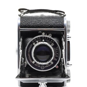 May include: A silver and black Telka II camera with a Demaria-Lapierre lens. The camera has a leather strap and a silver lever on the bottom.