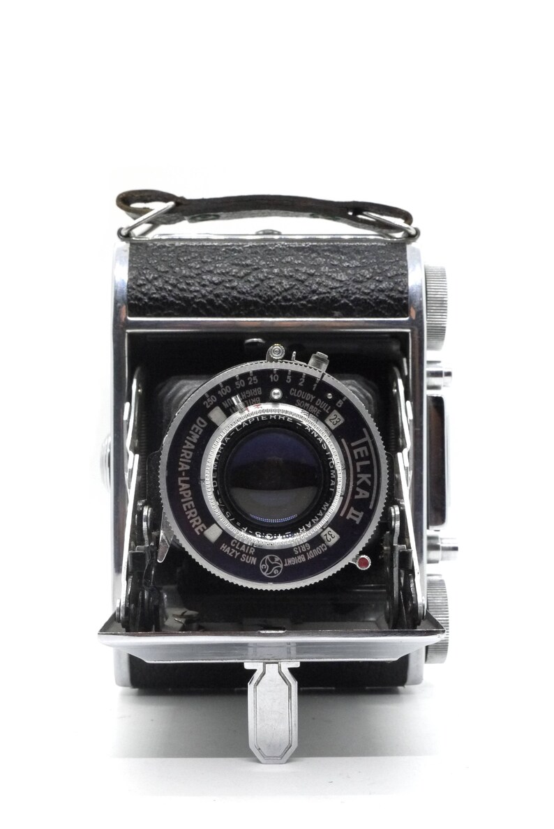 May include: A silver and black Telka II camera with a Demaria-Lapierre lens. The camera has a leather strap and a silver lever on the bottom.