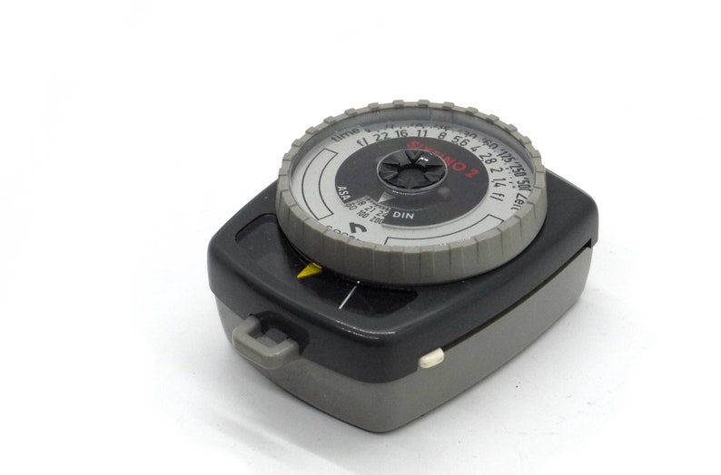 Puede incluir: Un fot&oacute;metro de mano gris y negro con un dial circular. El dial tiene marcas para el tiempo, ASA y DIN. El dial tambi&eacute;n tiene el texto "SEKONIC L" impreso en &eacute;l.