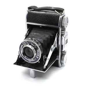 May include: A vintage Telka camera in silver and black with a leather covering on the top. The camera is folded up and the lens is visible.