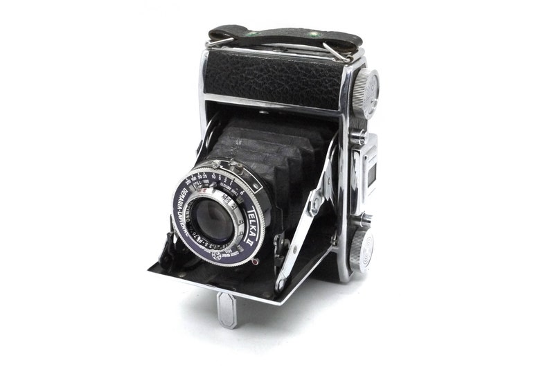 May include: A vintage Telka camera in silver and black with a leather covering on the top. The camera is folded up and the lens is visible.