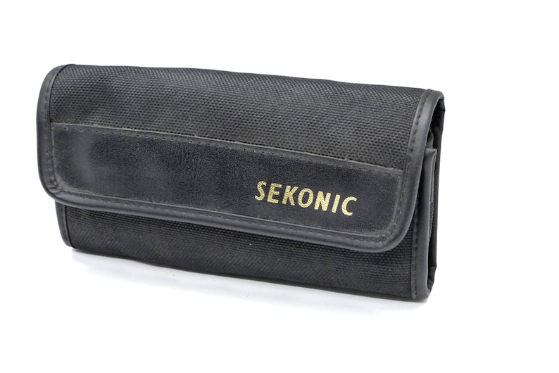 May include: Black canvas carrying case with a leather flap and the word "SEKONIC" printed in gold lettering.