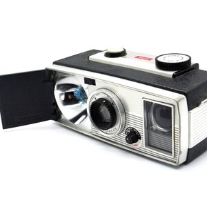 May include: A silver and black vintage camera with a built-in flash and a viewfinder. The camera has a black leatherette covering and a silver metal body. The camera has a red "WIND" button on the top.