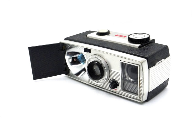 May include: A silver and black vintage camera with a built-in flash and a viewfinder. The camera has a black leatherette covering and a silver metal body. The camera has a red "WIND" button on the top.