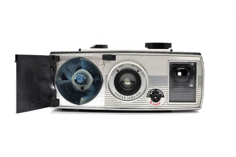 May include: A silver and black Kodak Instamatic camera with a built-in flash. The camera has a lens with a close-up and beyond setting. The camera has a built-in flash and a film advance lever.