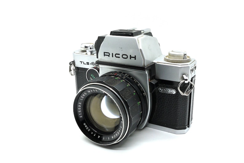 May include: A silver and black Ricoh TLS 400 SLR camera with a black 21/50.3 Ricoh Xenon lens attached. The camera has a black leatherette covering on the body.