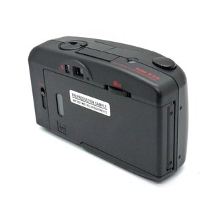 May include: A black camera with a red label that says "Preproduction Sample May Not Meet All Requirements". The camera has a silver button on the top and a black button on the right side. The camera is on a white background.