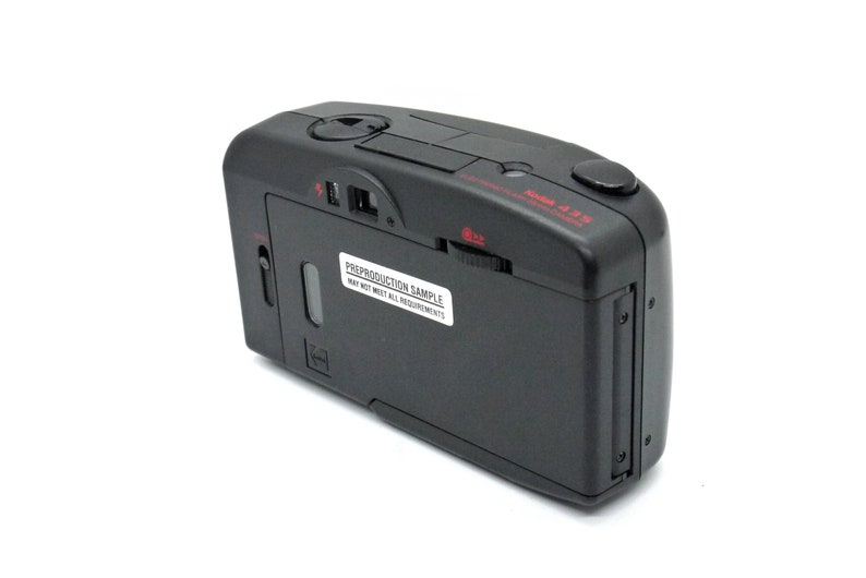 May include: A black camera with a red label that says "Preproduction Sample May Not Meet All Requirements". The camera has a silver button on the top and a black button on the right side. The camera is on a white background.