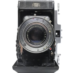 Puede incluir: Una c&aacute;mara vintage negra y plateada con una lente etiquetada "Tovar-Anastigmat 1:4.5 f=105mm 22" y "ZEISS IKON" en la parte superior.