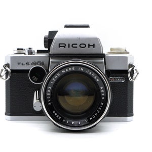 May include: A silver and black Ricoh TLS 401 35mm film camera with a Rikenon 55mm f/1.9 lens. The camera has a black leatherette covering and a silver metal body.