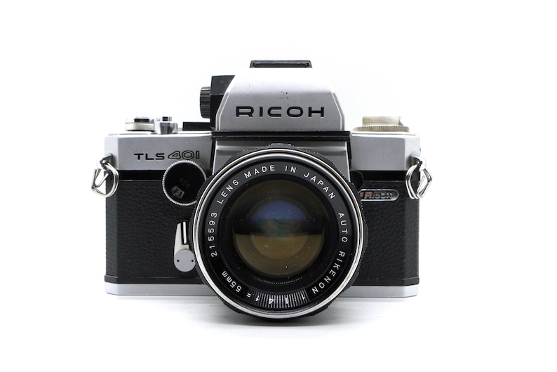 May include: A silver and black Ricoh TLS 401 35mm film camera with a Rikenon 55mm f/1.9 lens. The camera has a black leatherette covering and a silver metal body.