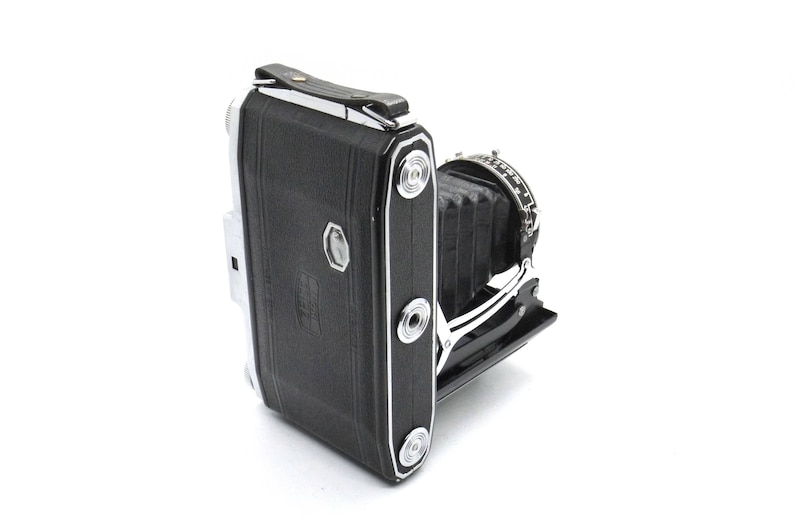 Puede incluir: Una c&aacute;mara plegable negra y plateada con fuelle y cubierta de cuero. La c&aacute;mara tiene un acabado cromado y una correa de cuero.