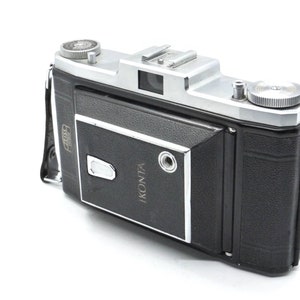 Puede incluir: Una c&aacute;mara Ikonta plateada y negra con una cubierta de cuero. La c&aacute;mara tiene una lente plateada y una correa de cuero negra.