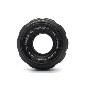 May include: A black camera lens with the text "LENS MADE IN JAPAN EL-NIKKOR 1:5.6 f=105mm Nippon Kogaku Japan No.326333" printed around the edge.