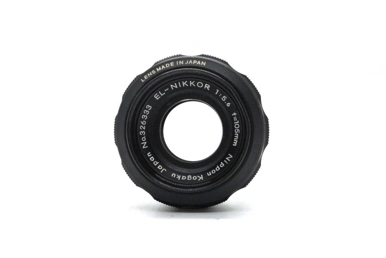 May include: A black camera lens with the text "LENS MADE IN JAPAN EL-NIKKOR 1:5.6 f=105mm Nippon Kogaku Japan No.326333" printed around the edge.