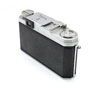 Op de afbeelding: Een zilveren en zwarte vintage camera met de naam "Kowa" erop gedrukt. De camera heeft een zwarte lederen bekleding en een zilveren metalen behuizing.