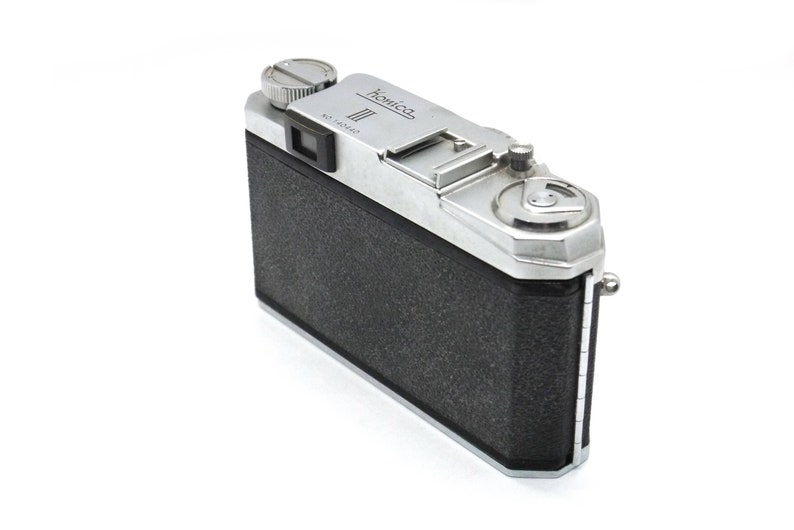 Op de afbeelding: Een zilveren en zwarte vintage camera met de naam "Kowa" erop gedrukt. De camera heeft een zwarte lederen bekleding en een zilveren metalen behuizing.