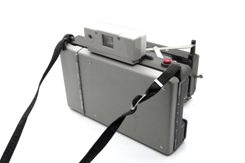 May include: A gray Polaroid camera with a black strap. The camera has a red button on the side and a white flash unit on top.