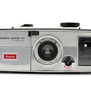 May include: A silver Kodak Brownie Super 27 camera with a black lens and a flash setting. The camera has a red Kodak logo and the text "BROWNIE SUPER 27 CAMERA" on the front.