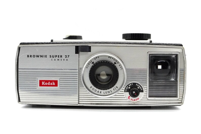 May include: A silver Kodak Brownie Super 27 camera with a black lens and a flash setting. The camera has a red Kodak logo and the text "BROWNIE SUPER 27 CAMERA" on the front.