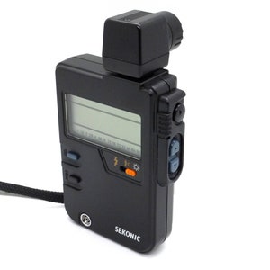 May include: A black Sekonic light meter with a digital display and a built-in flash sensor. The meter has a black strap and a small, black, square-shaped flash head.