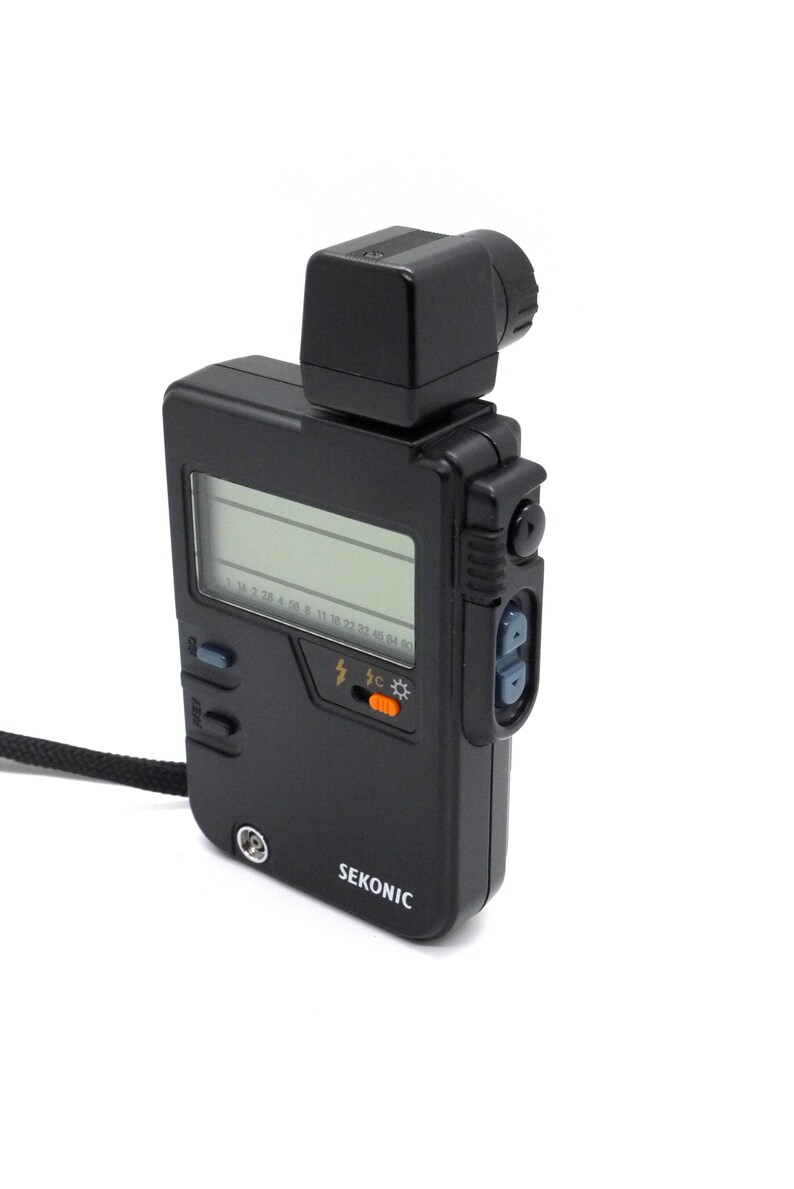 May include: A black Sekonic light meter with a digital display and a built-in flash sensor. The meter has a black strap and a small, black, square-shaped flash head.