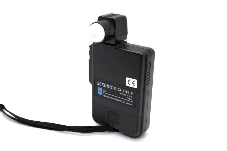 May include: A black Sekonic Digi Lite F light meter with a white dome on the top. The meter has a black strap and the text "SEKONIC DIGI LITE F MODEL L-328 C340 SEKONIC CORPORATION JAPAN" is printed on the side.