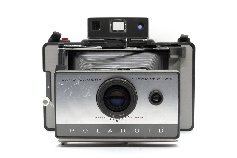 May include: A silver and black Polaroid Land Camera Automatic 103 with a dark lens and a light adjustment dial. The camera has a black strap and a red button.