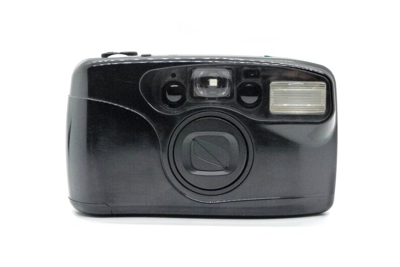 May include: A black point-and-shoot film camera with a built-in flash. The camera has a lens, flash, and viewfinder. The camera is on a white background. The camera is a vintage style.