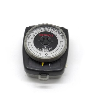 Puede incluir: Un fot&oacute;metro Gossen Sixtrino 2 negro y gris con una esfera blanca y una aguja que apunta hacia la derecha. La esfera tiene marcas para el tiempo, la apertura, la ASA y el DIN.